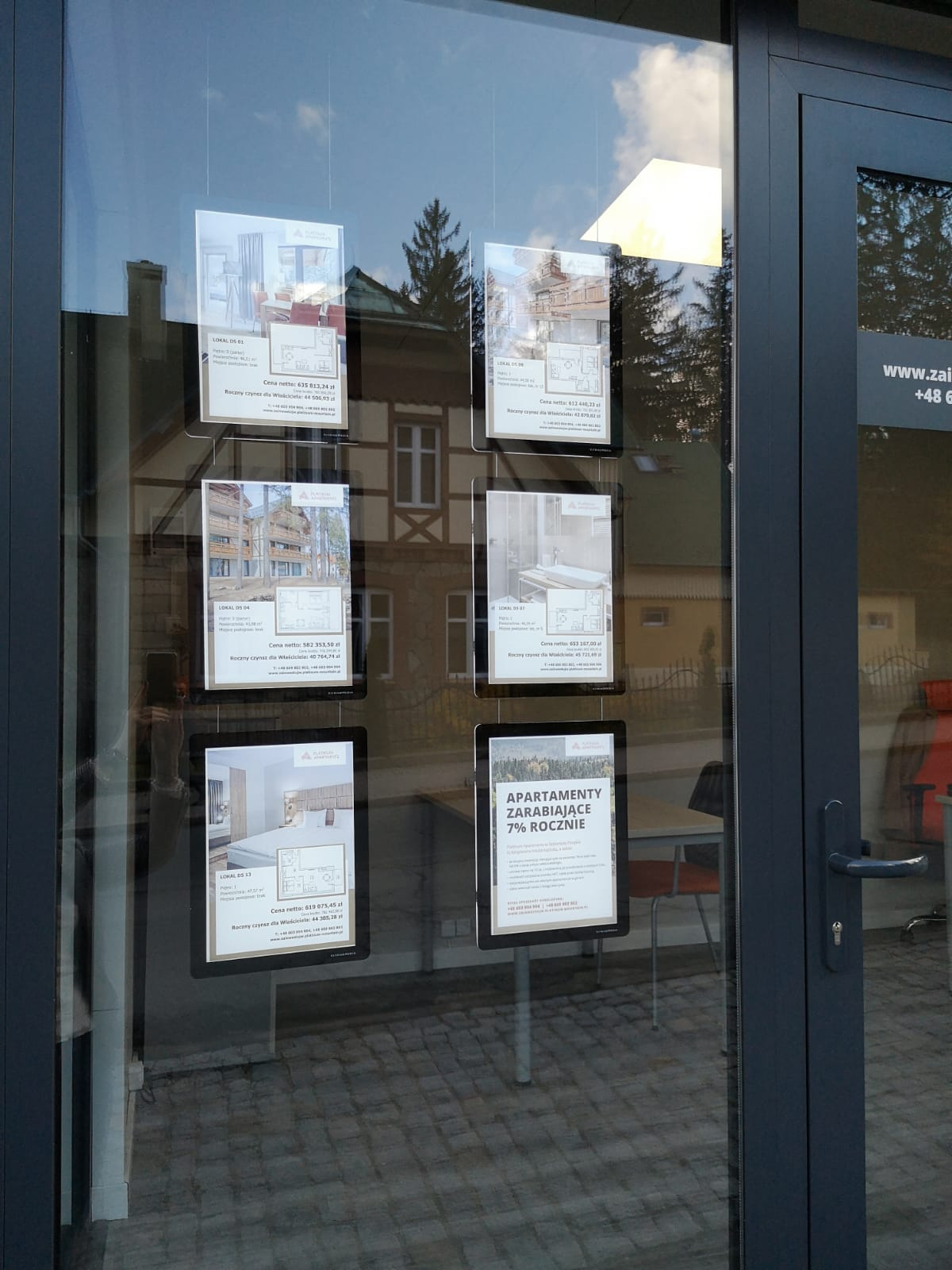 Witryna biura sprzedaży wykorzystująca system linkowy LED do prezentacji materiałów reklamowych. Oferty mieszkań z opisami, zdjęciami i rzutami pomieszczeń są umieszczone w podświetlanych kieszeniach akrylowych zawieszonych na stalowych linkach. Zdjęcie wykonano w dzień, dlatego efekt podświetlenia jest mniej widoczny.