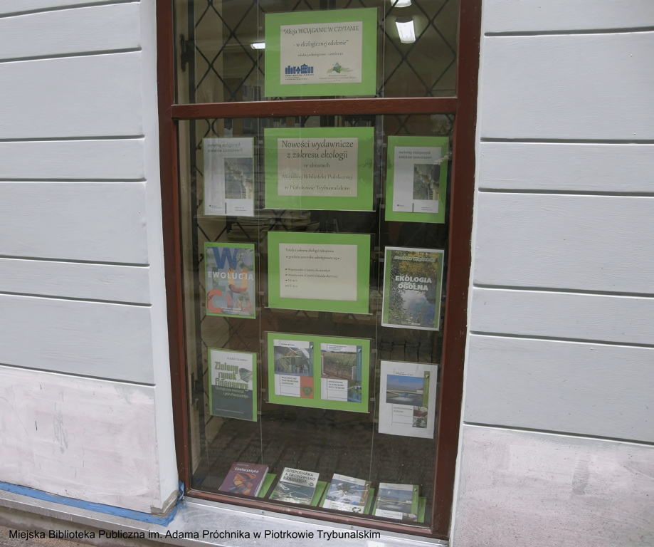 Witryna biblioteki miejskiej z ekspozycją materiałów informacyjnych i promocyjnych o tematyce ekologicznej, wykorzystująca tradycyjny system linkowy z przezroczystymi kieszeniami akrylowymi. Widoczne są plakaty i ulotki związane z ochroną środowiska oraz nowościami wydawniczymi.