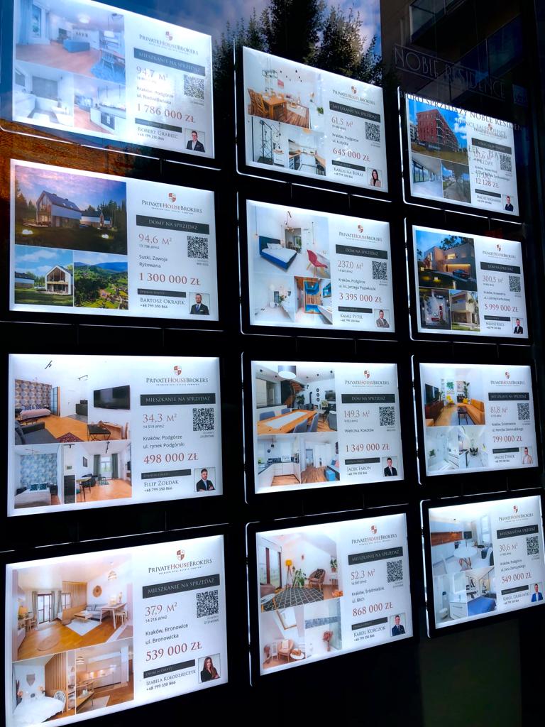 djęcie prezentuje system linkowy LED wykorzystywany do ekspozycji ofert sprzedaży nieruchomości w witrynie biura nieruchomości. Oferty umieszczone są w akrylowych, podświetlanych kieszeniach, które wyraźnie prezentują zdjęcia, opisy oraz ceny oferowanych mieszkań i domów. Całość zapewnia elegancki wygląd i dobrą widoczność nawet przy słabszym oświetleniu.