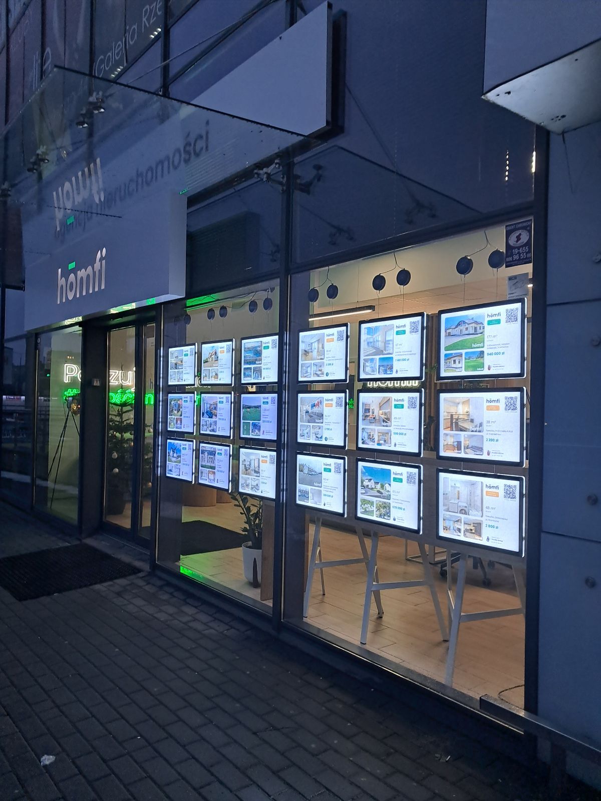 djęcie prezentuje system linkowy LED wykorzystywany do ekspozycji ofert sprzedaży nieruchomości w witrynie biura nieruchomości. Oferty umieszczone są w akrylowych, podświetlanych kieszeniach, które wyraźnie prezentują zdjęcia, opisy oraz ceny oferowanych mieszkań i domów. Całość zapewnia elegancki wygląd i dobrą widoczność nawet przy słabszym oświetleniu.