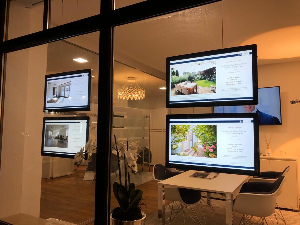Zdjęcie przedstawia nowoczesny system linkowy LCD zastosowany w witrynie biura nieruchomości. Ekrany LCD zawieszone na stalowych linkach prezentują cyfrowe oferty mieszkań, domów oraz ogrodów. Ekspozycja jest dobrze widoczna dzięki jasnym ekranom i profesjonalnie komponuje się z eleganckim wystrojem wnętrza biura.